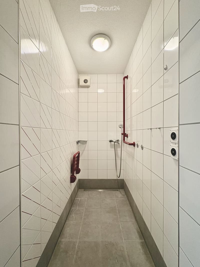 Modern shower area with white tiled walls and floor, a single showerhead, a faucet, and red handrails for support.