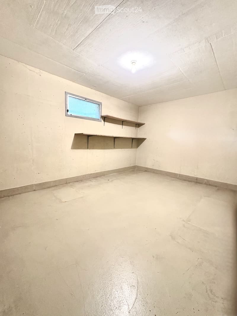 Empty room with concrete floor, beige walls, and two metal shelves mounted on the wall. A small window with a metal frame is visible on one wall.