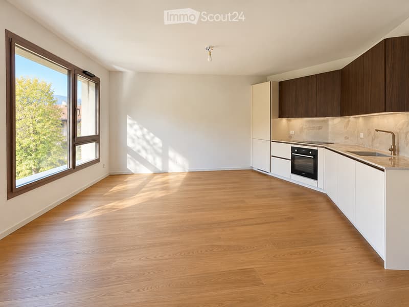 Empty kitchen with wooden flooring, white cabinets, and large windows allowing natural light. No furniture or appliances are present.