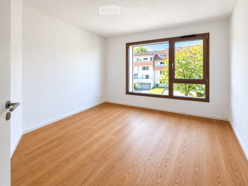 Empty room with light wooden flooring and white walls, featuring a large window with brown frame overlooking a residential area.