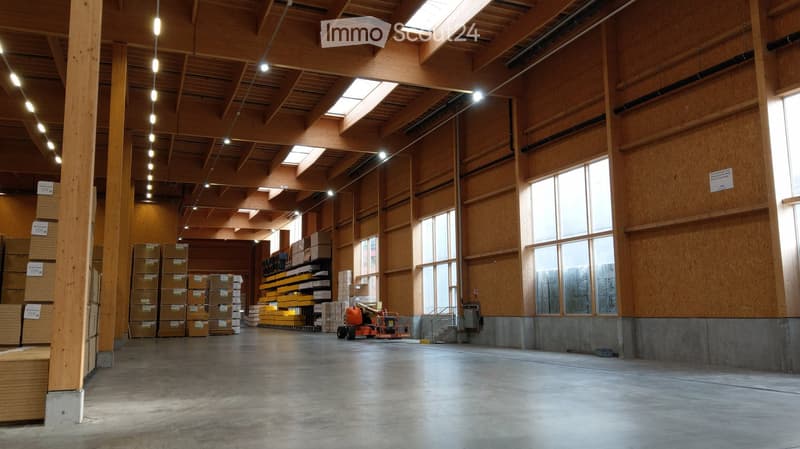 Large industrial-style warehouse with exposed wooden beams, concrete floor, large windows, and stacks of wooden pallets and storage shelves.