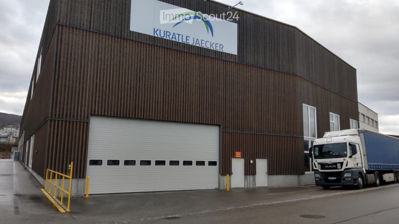 Large industrial building with a corrugated metal facade, large garage door, and a sign reading "KURATLE JAECKER". A semi-truck is parked in front.