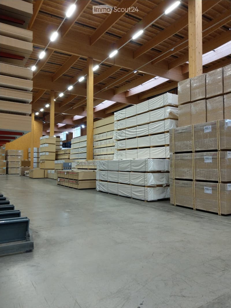 Large industrial warehouse with wooden beams, concrete floor, and stacks of wooden panels and cardboard boxes.