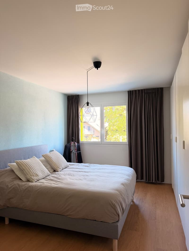 A modern bedroom with a large bed, pillows, and a hanging geometric lamp. The room features wooden flooring, large windows with dark curtains, and a wardrobe with a jacket hanging on it.