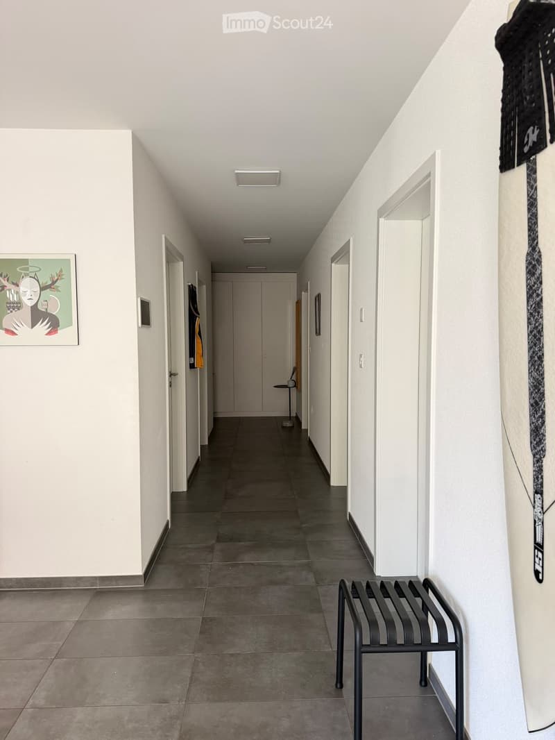 A long, narrow hallway with white walls, tiled floor, and several doors leading to rooms. A bench is placed on the right side, and a framed artwork hangs on the left wall.