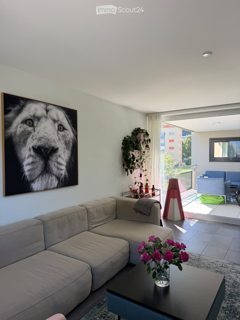 Modern living room with a beige sectional sofa, coffee table with a vase of pink flowers, side table with bottles, and a large picture frame on the wall. The room has large windows leading to a balcony with outdoor seating.