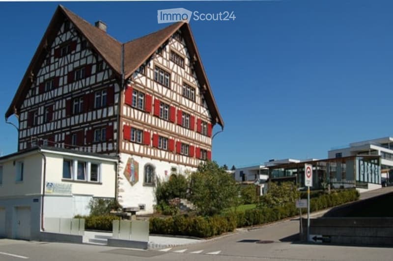 Traditional Swiss-style building with a steeply pitched roof, exposed wooden beams, and red shutters. The structure is two stories with multiple windows and a small balcony on the upper level.