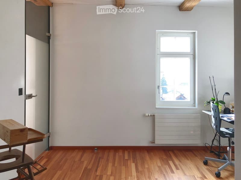 A small, unfurnished room with wooden flooring, a window, a radiator, a small table with a wooden box, a chair, a desk with a lamp, and a plant.