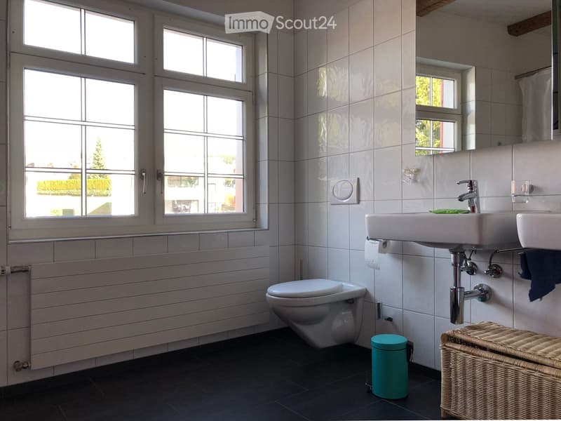 Modern bathroom with white tiles, a toilet, double sink, mirror, and a wicker basket on the floor.