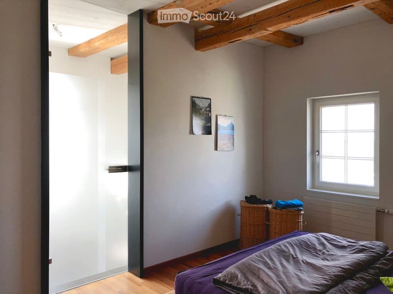 Bedroom with wooden floor, exposed wooden beams, a bed with purple bedding, two wicker baskets, and two picture frames on the wall.