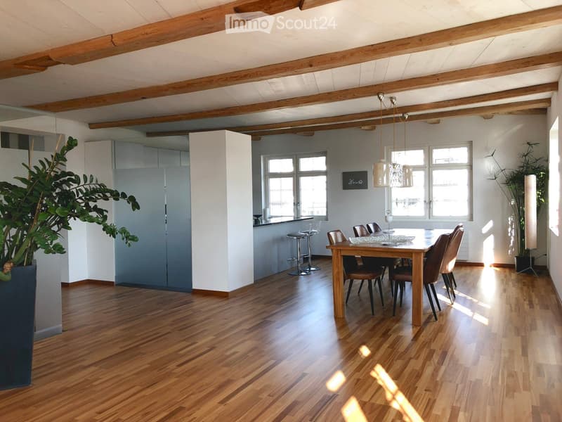 Open-plan living and dining area with wooden floors, exposed wooden beams, large windows, a dining table with chairs, bar stools, and pendant lighting.