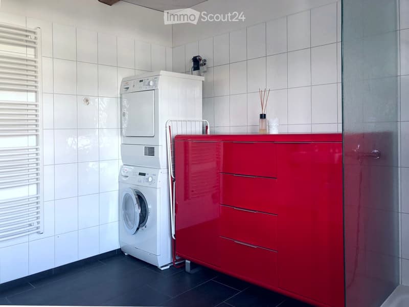 Modern bathroom with white tiled walls, a stacked washing machine and dryer, red cabinet, towel rack, and decorative items on the counter.
