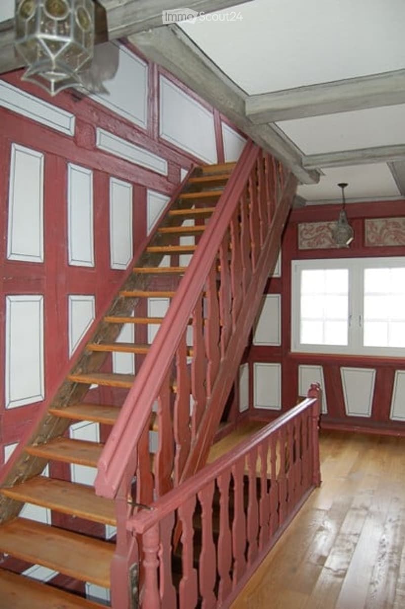 A wooden staircase with red-painted railings and white paneling on the walls, leading up to a higher level. The hallway has wooden flooring and a chandelier hanging from the ceiling.