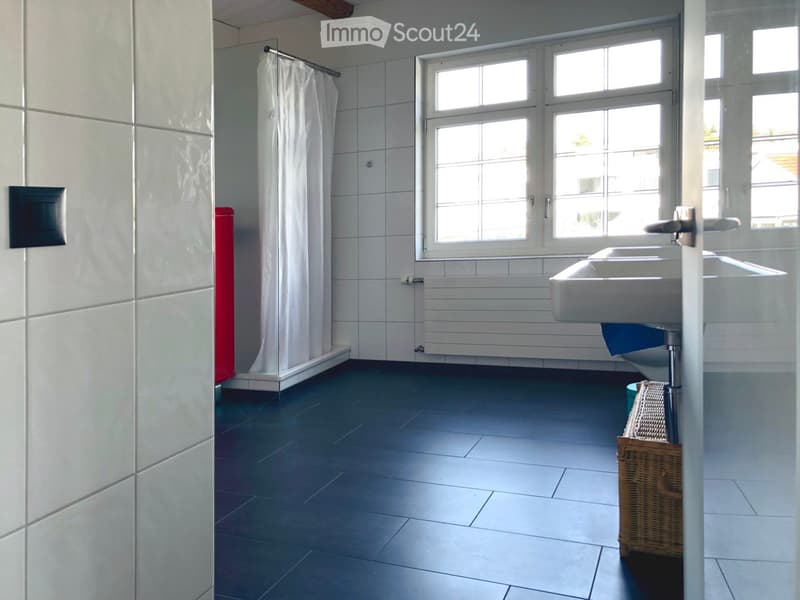 Modern bathroom with white tiled walls, dark grey tiled floor, large window, white sink, shower area with white curtain, and a wicker basket on the floor.
