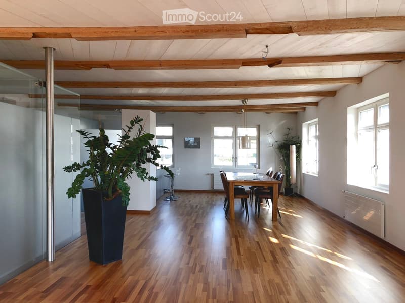 Open-plan dining area with wooden flooring, exposed wooden beams, large windows, dining table with chairs, and a tall potted plant.