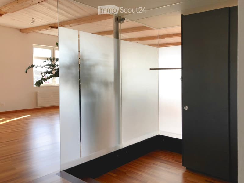 An unfurnished room with wooden flooring, white walls, and a large window with a plant visible outside. The room features a modern glass partition and a black door.