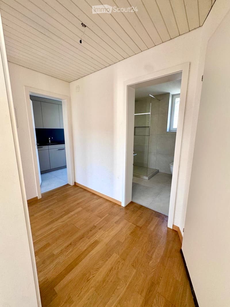 Empty hallway with light wooden flooring and white walls, leading to a kitchen and a bathroom with a glass-enclosed shower and a toilet.