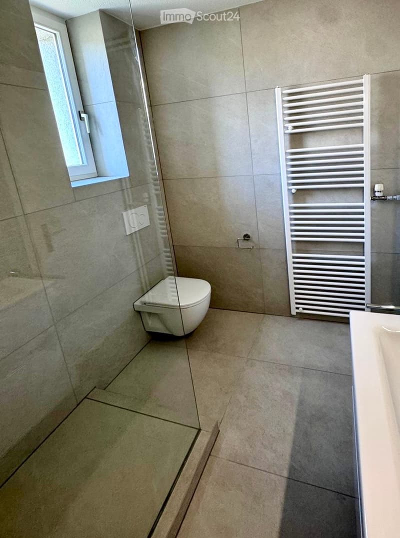 Modern bathroom with beige tiles, a toilet, a radiator, and a sink. The space is clean and well-lit with a window providing natural light.