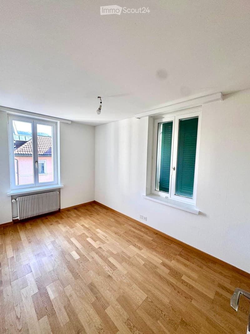 Empty room with light wooden flooring, white walls, and two windows with shutters. A radiator is visible under one window, and a single light fixture hangs from the ceiling.