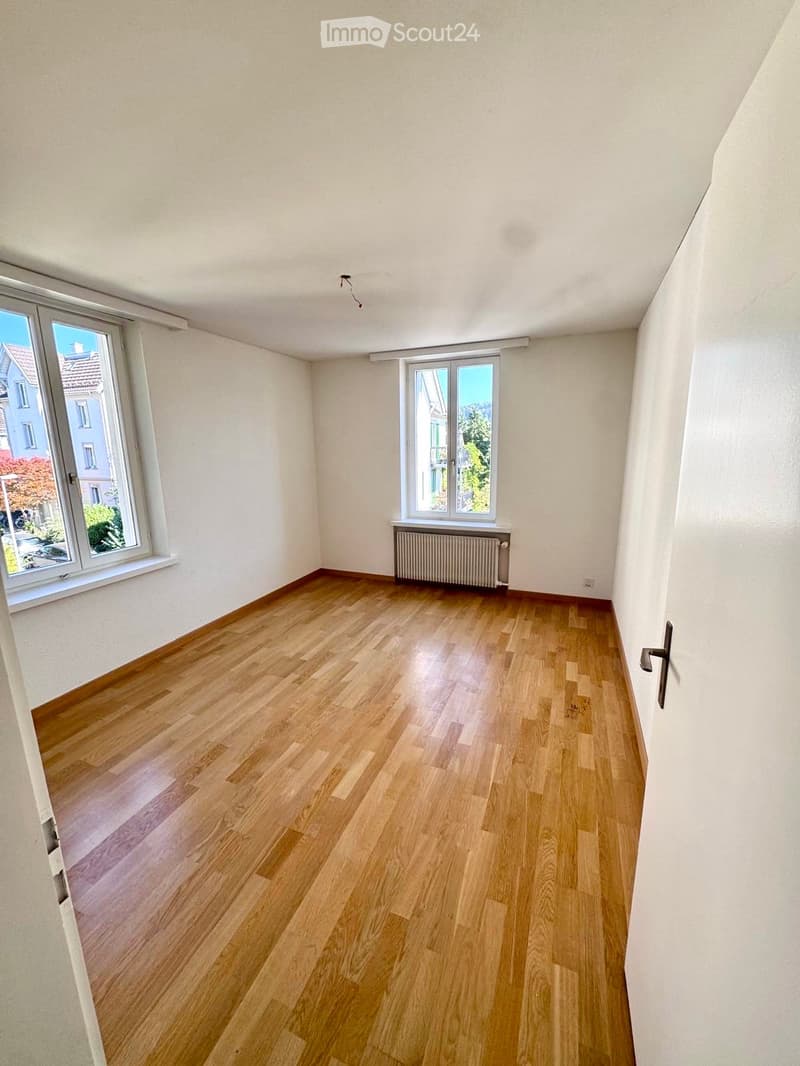 Empty room with wooden flooring, white walls, and two windows offering a view of the street and neighboring buildings.