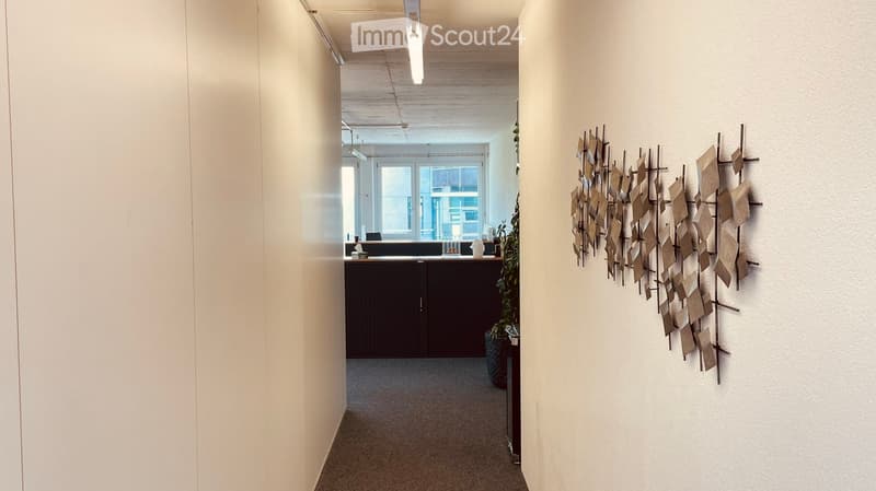 A modern hallway with light-colored walls, a geometric wall art piece, and a small reception area visible at the end.