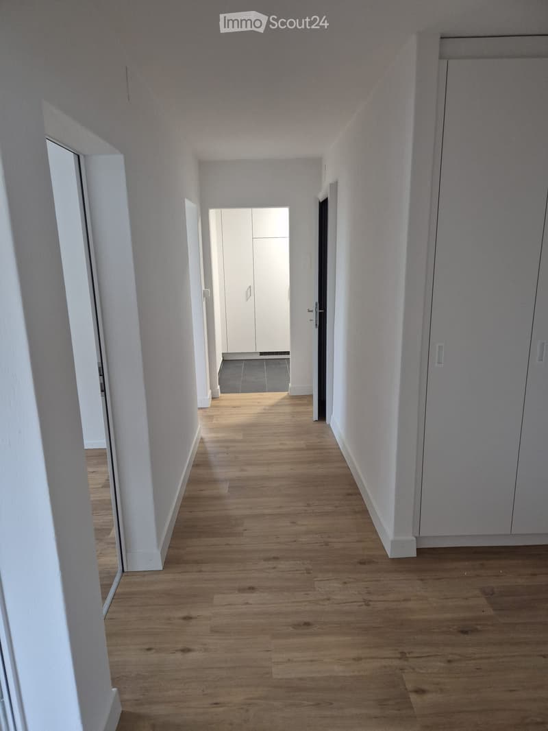 Hallway with white walls, wooden flooring, white built-in cabinets, and an open door to the kitchen.