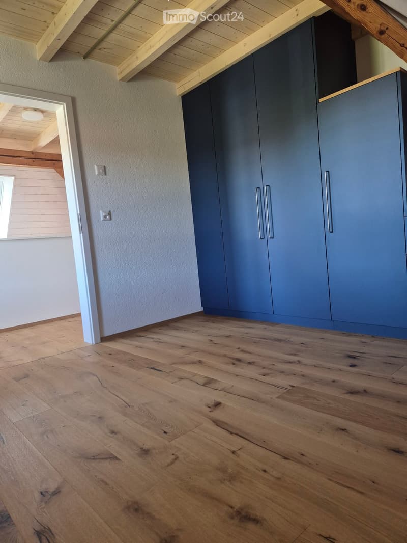 Empty room with wooden flooring, light-colored walls, and modern grey cabinets with silver handles. The ceiling features exposed wooden beams.