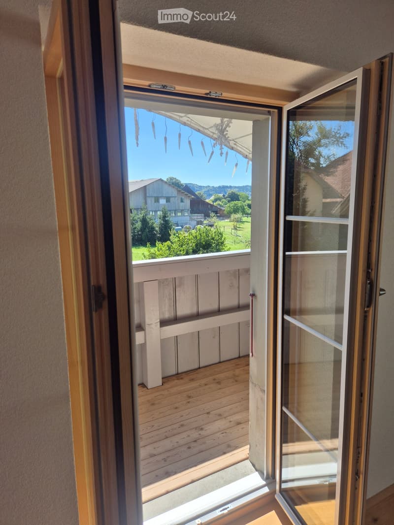 Open sliding door leads to a wooden balcony with a view of a green landscape and houses.
