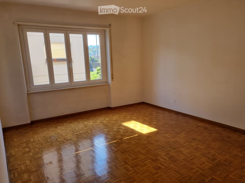 Empty room with wooden parquet flooring and large windows allowing natural light to enter.