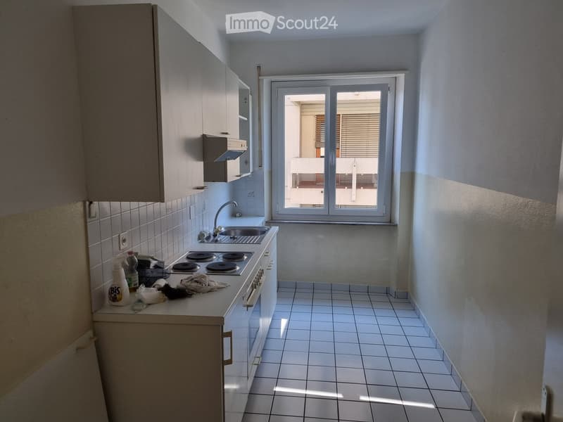 Modern kitchen with white tiled walls and floor, fitted cabinets, a sink, a stove, and a window leading to a balcony.