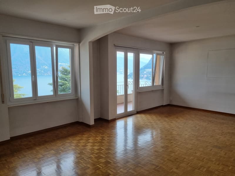 Empty room with wooden flooring, large windows offering a scenic lake view, and a sliding glass door leading to a balcony.