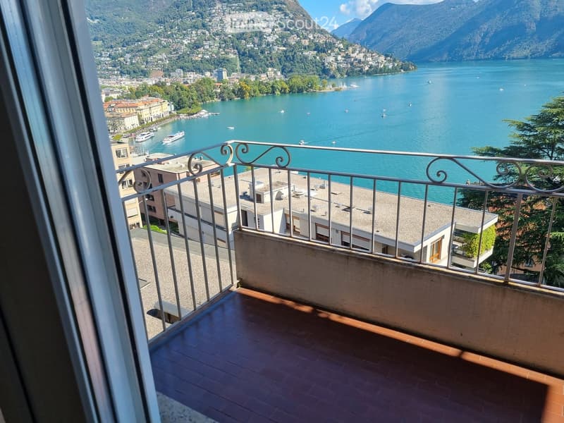 Balcony with wooden flooring and metal railing, overlooking a scenic lake and town with boats and mountains in the background.
