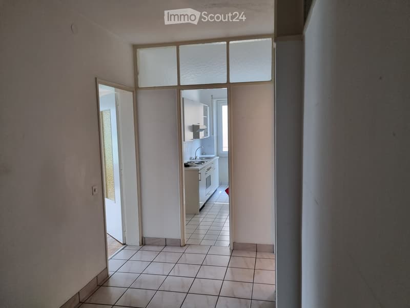 Empty hallway with tiled floor, leading to a small kitchen area with white cabinets, sink, and stove.
