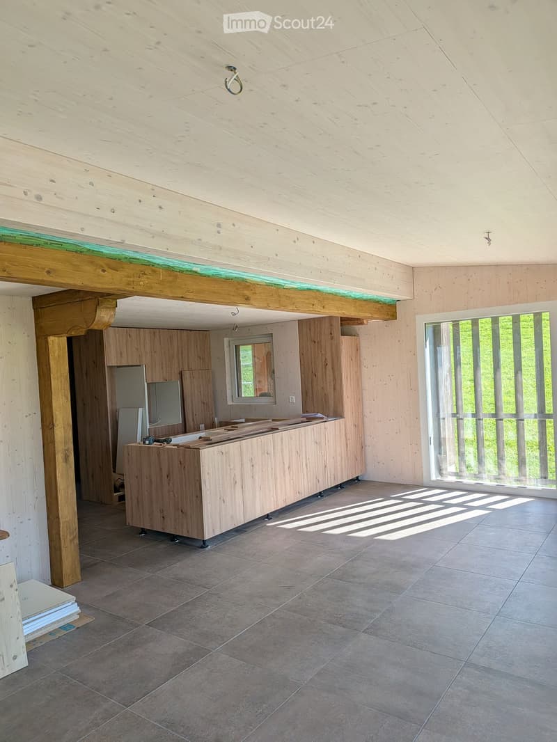 Empty room with wooden ceiling beams, tiled floor, large window, and unfinished wooden cabinetry.