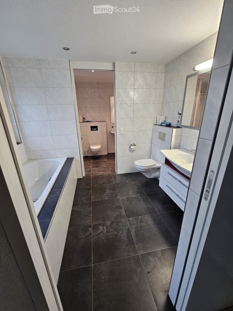 Modern bathroom with dark tiled floor, white bathtub, two toilets, sink with cabinet, and mirror.