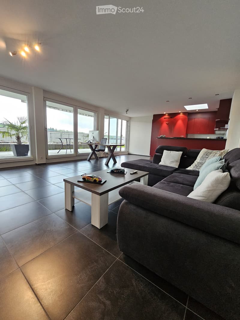 Modern living room with a grey sectional sofa, coffee table, laptop, and large windows leading to a balcony.