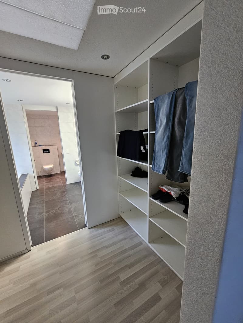 Empty room with wooden flooring, white built-in closet with empty shelves, and an adjacent bathroom with a toilet and tiled walls.