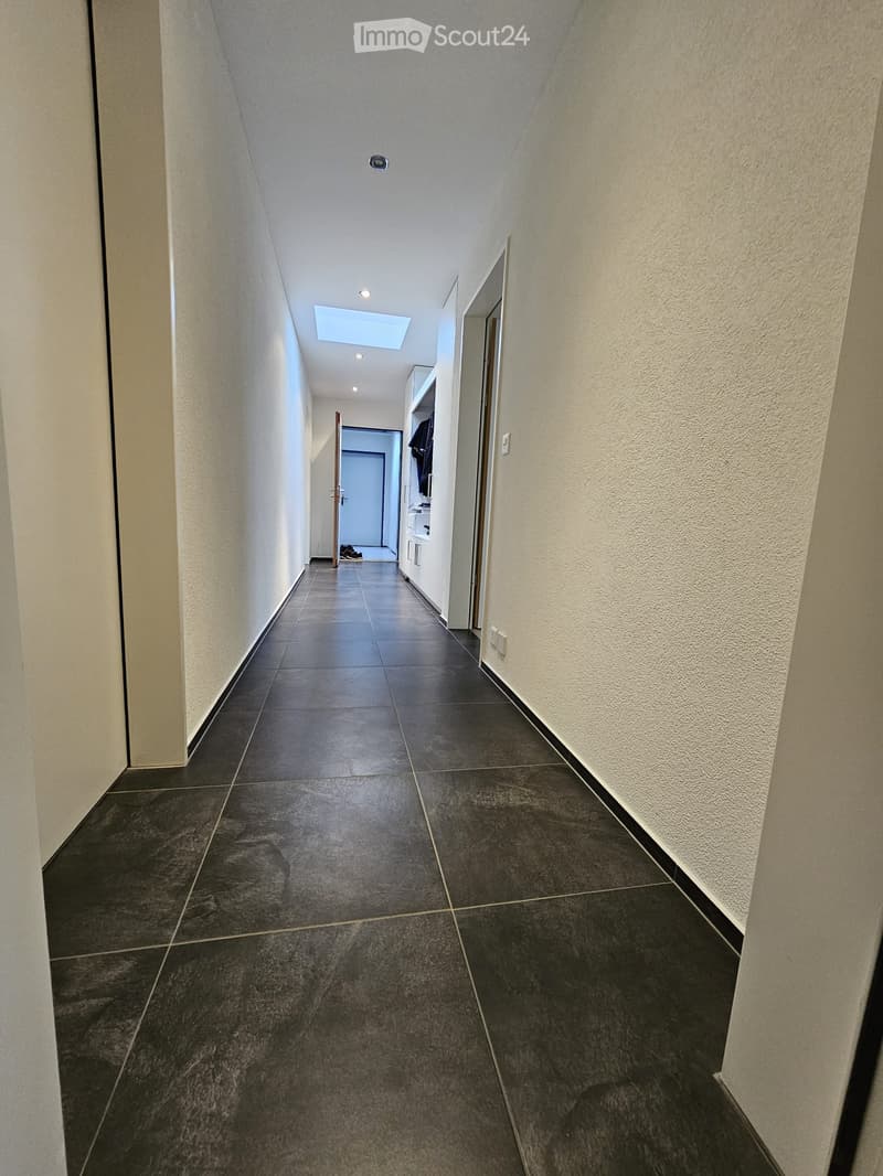 Clean, modern hallway with dark tiled flooring, white walls, and recessed lighting. Features a small skylight and a partially visible closet with clothes hanging inside.