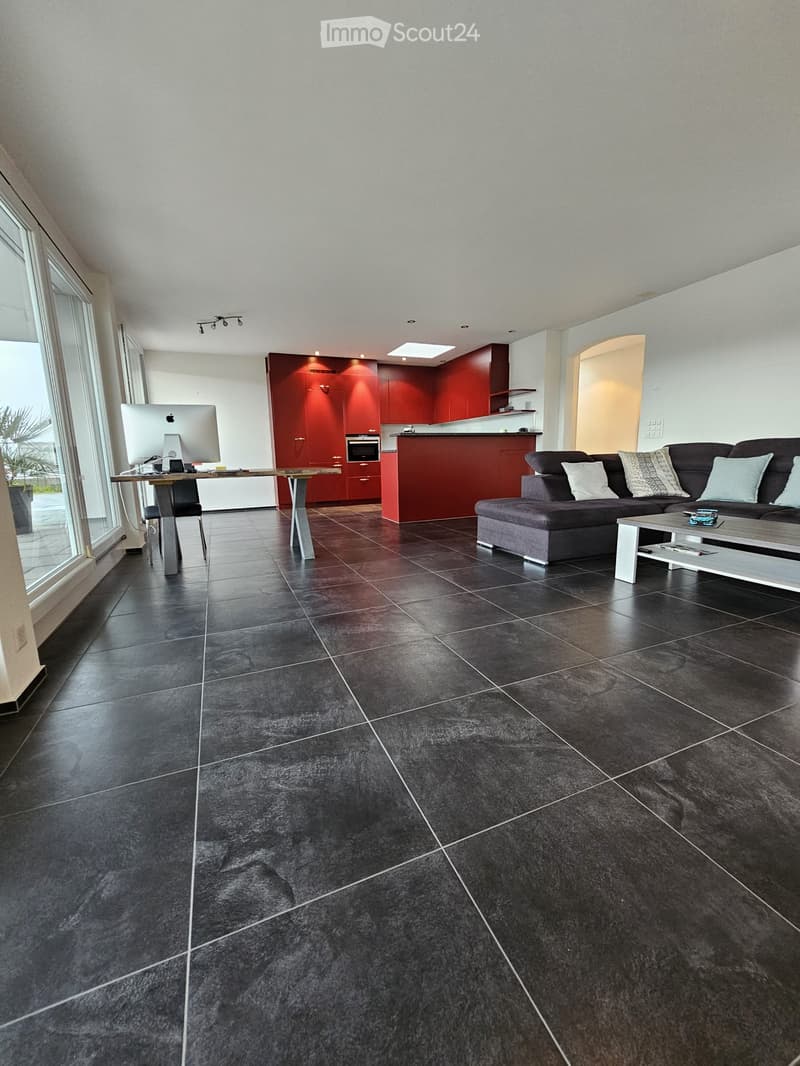 Modern living room with dark tiled floor, grey sectional sofa, coffee table, computer desk, and red kitchen area with cabinets and oven.