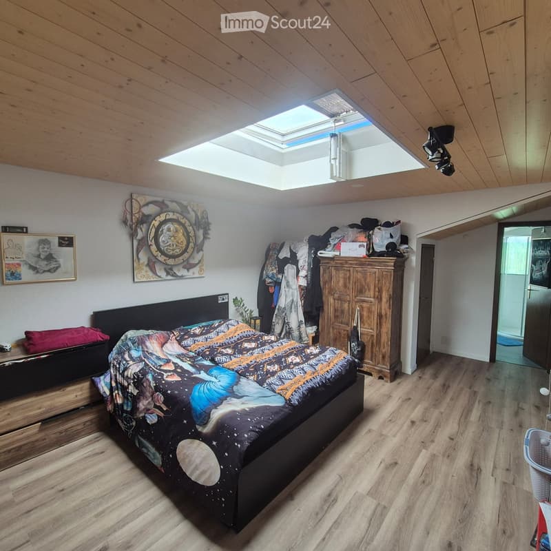 Bedroom with wooden floors, slanted wooden ceiling, skylight window, bed with colorful space-themed bedding, nightstand, wooden cabinet, framed picture, clothing rack, and small plant.