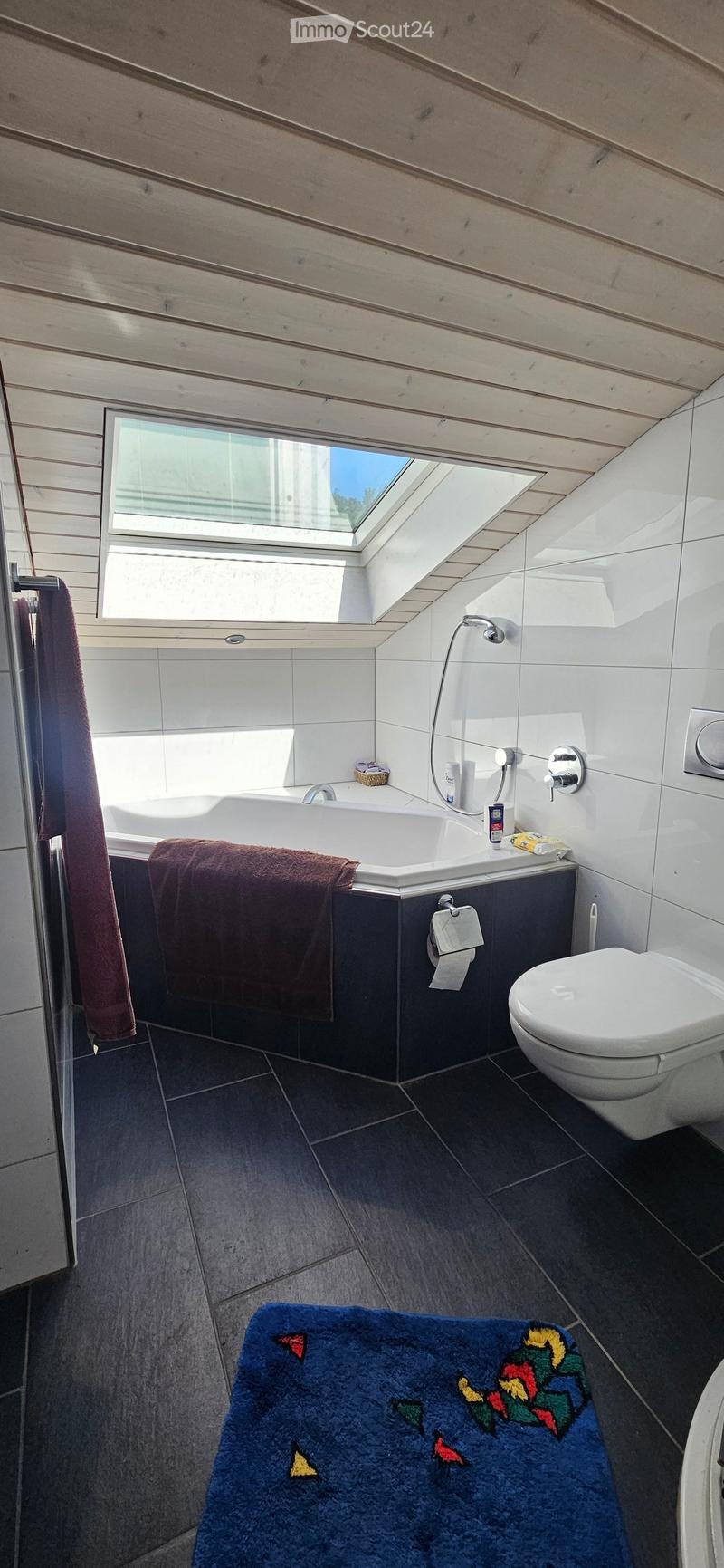 Modern bathroom with a bathtub, toilet, towel, and a blue rug with a colorful design. Features a skylight window and sleek fixtures.