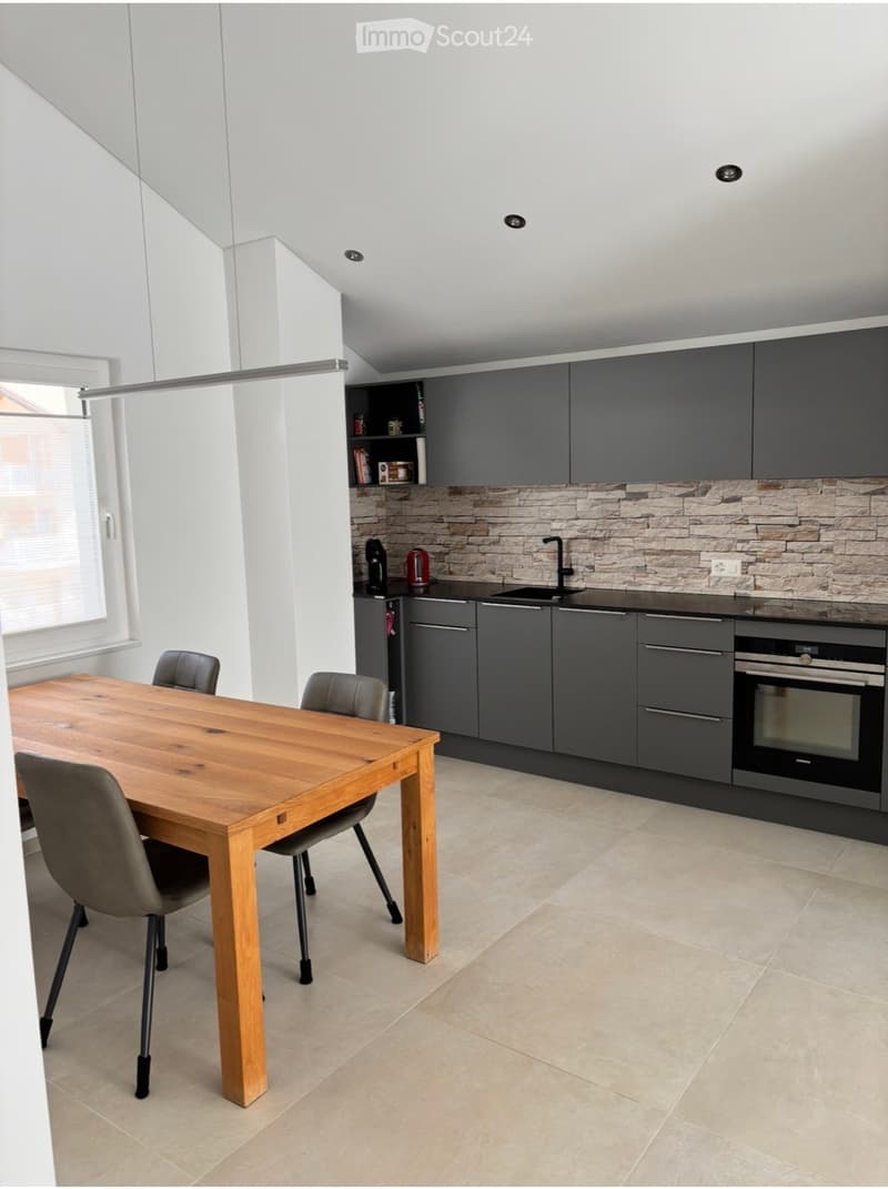 Modern kitchen with grey cabinets, stone backsplash, wooden dining table, and four chairs.