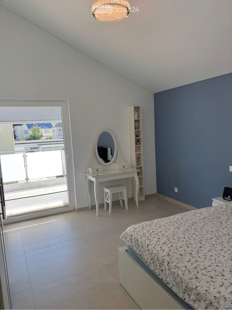 Bright bedroom with a bed, vanity table, mirror, and shelf. Large window with balcony access and a chandelier hanging from the ceiling.