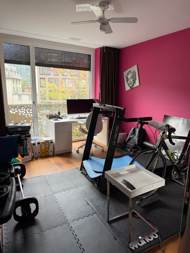 A multi-purpose room with a desk, exercise bike, bicycle, and various office supplies. The room features pink walls, large windows with blinds, and a ceiling fan.