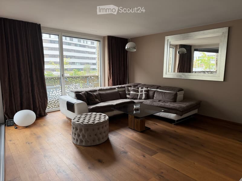Modern living room with a sectional sofa, coffee table, ottoman, floor lamp, and large mirror. The room features wooden flooring and large windows with curtains.
