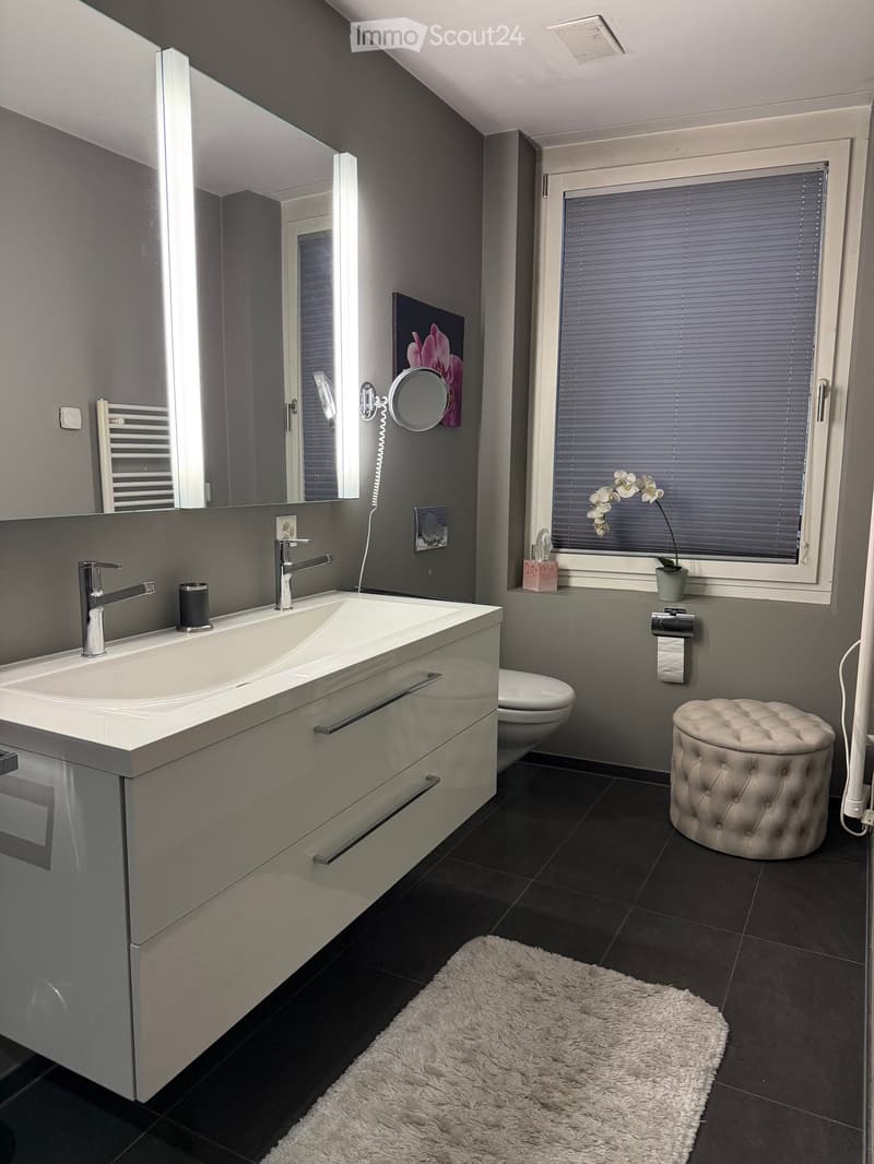Modern bathroom with a double sink, large mirror, toilet, tufted stool, and a fluffy white rug on the floor. A window with blinds and a small potted plant is visible.