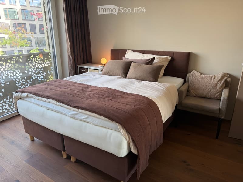 Cozy bedroom with a large bed, nightstand, lamp, and a small chair, featuring wooden flooring and large windows with a view of the city.