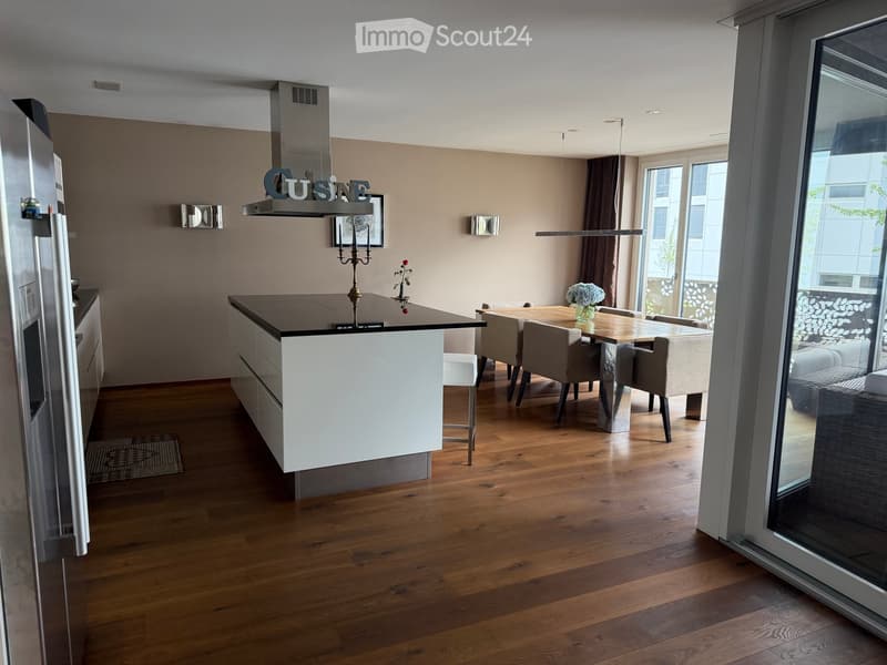 Modern kitchen and dining area with wooden flooring, white cabinets, stainless steel appliances, and a central island. A dining table with chairs is placed near large windows offering a view of the outside.