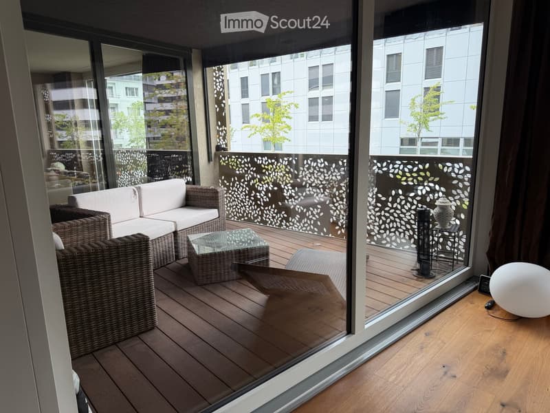 Balcony with wooden decking, wicker furniture, glass coffee table, decorative vase, and a lamp visible through large glass doors.