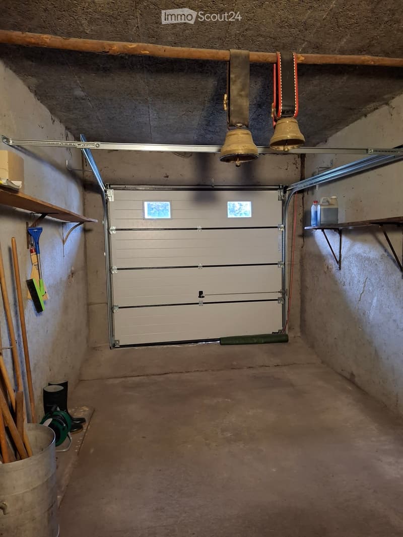 Empty garage with concrete floor, metal garage door, shelves, and some stored items like brooms and containers.